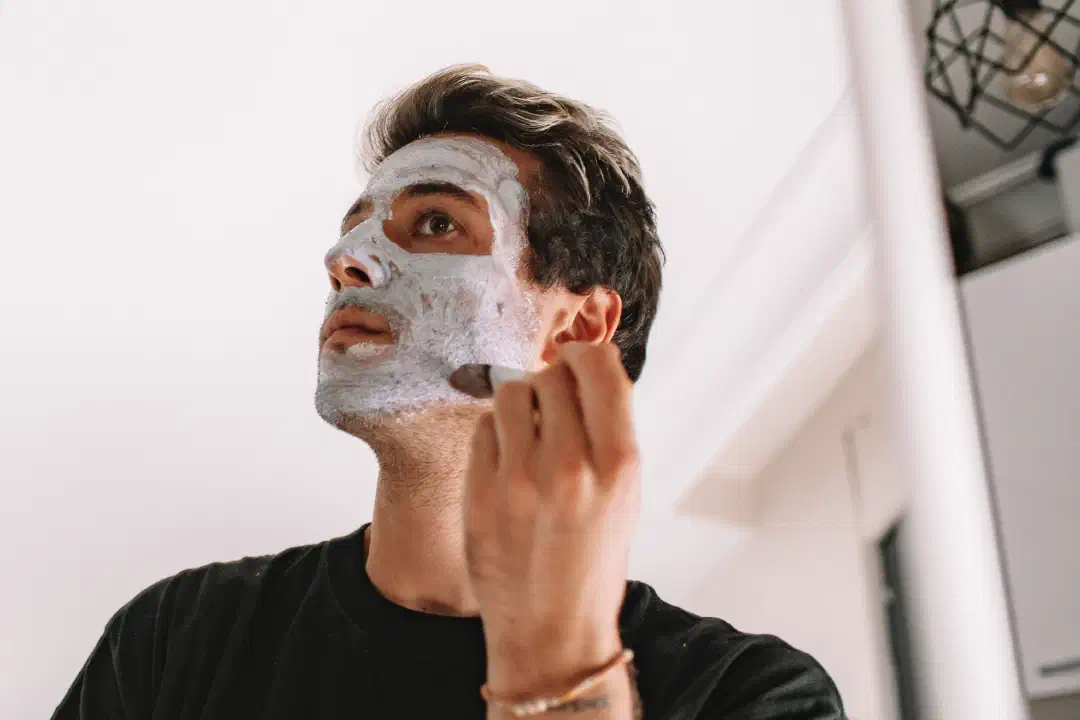 An adult man applying a facial mask while looking in a mirror at home; photo by ROMAN ODINTSOV